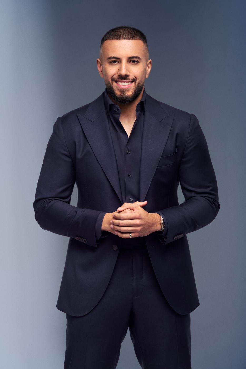 Man in a black suit with hands clasped, smiling