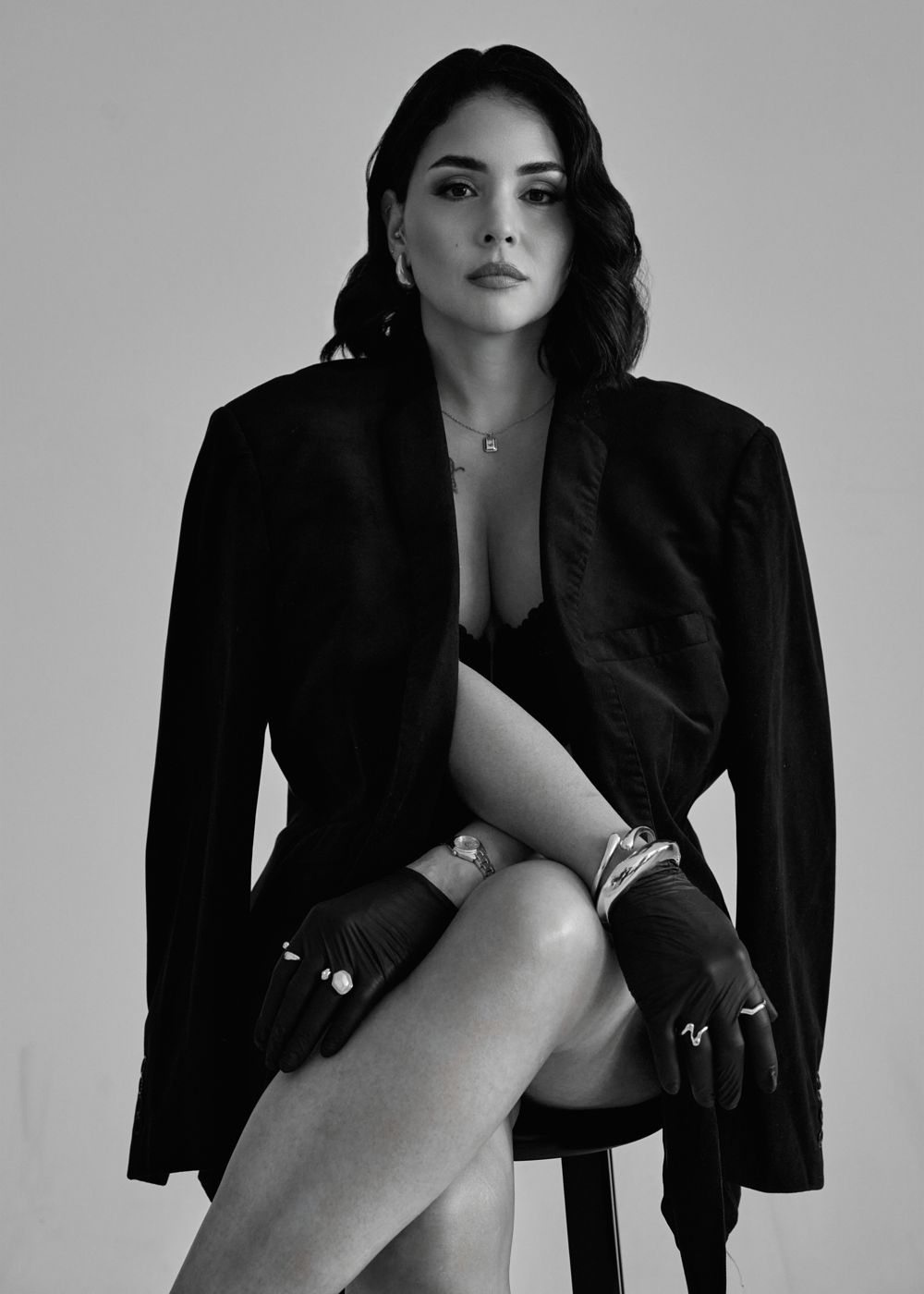 Black and white portrait of a woman in an oversized blazer and gloves on a stool