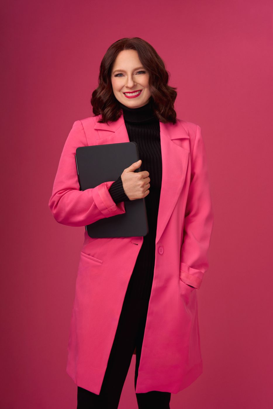 Smiling woman in a pink coat holding a tablet against a magenta backdrop