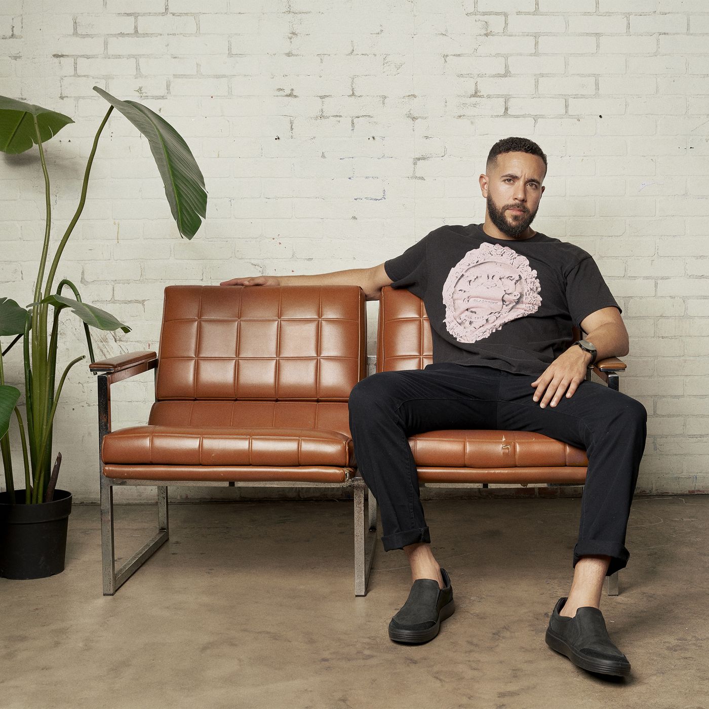 Man seated on a leather sofa next to a plant in a white brick space