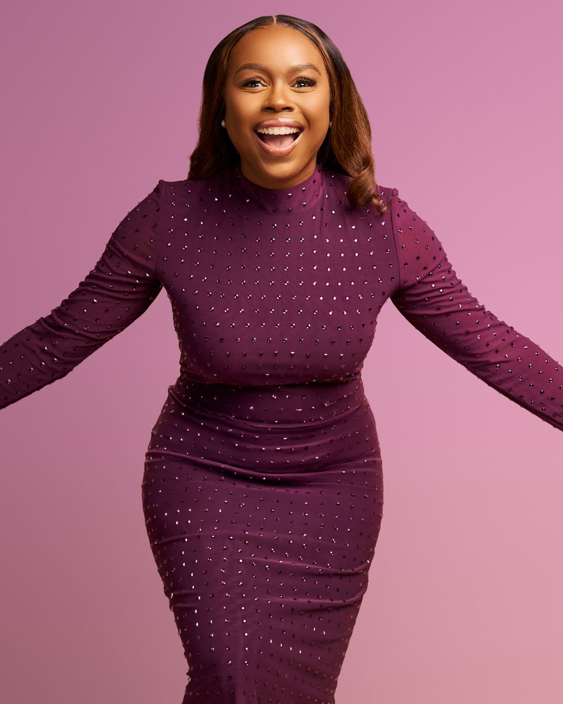 Smiling woman in a plum dress with arms open against a pink backdrop