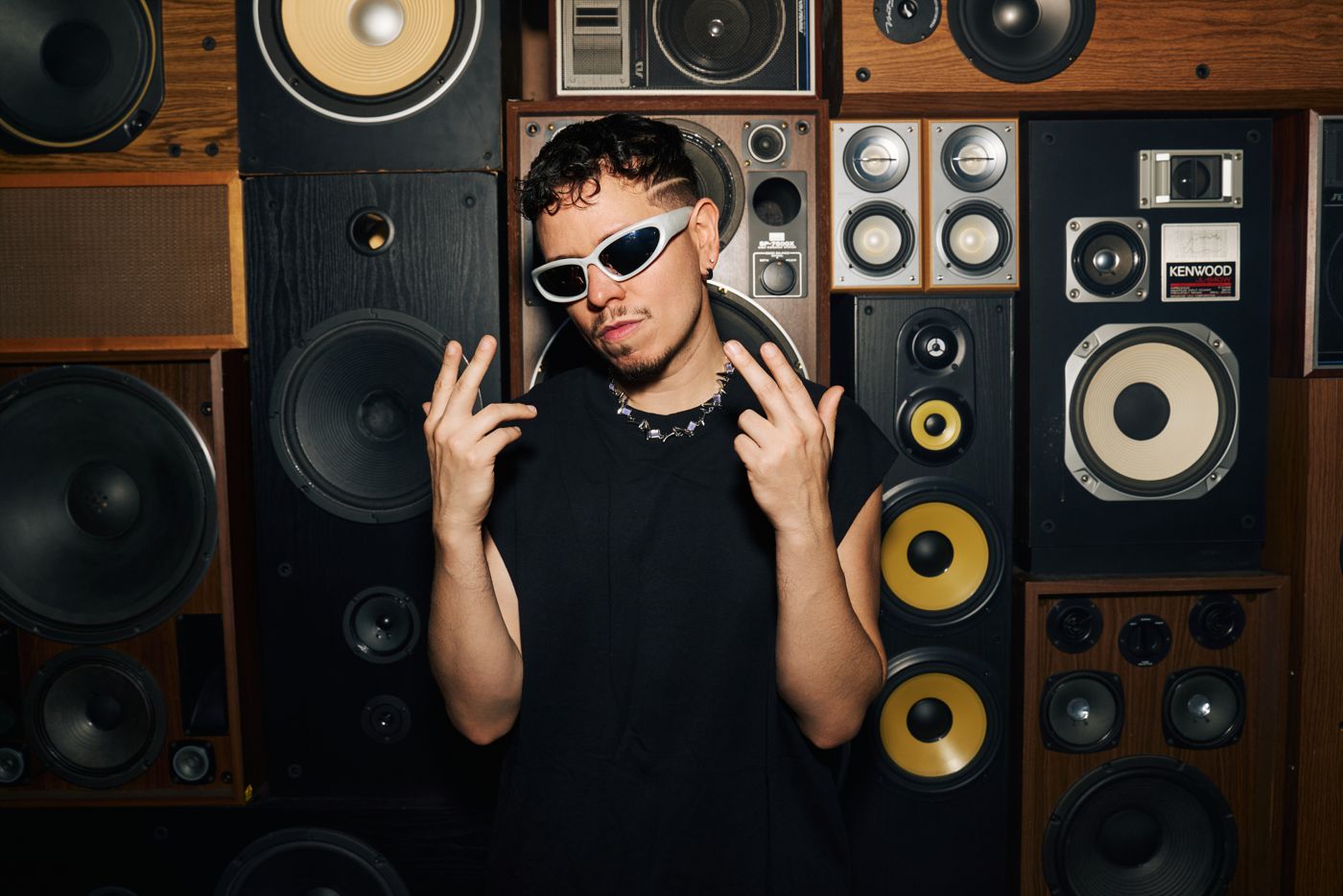 Artist in sunglasses posing in front of a wall of speakers